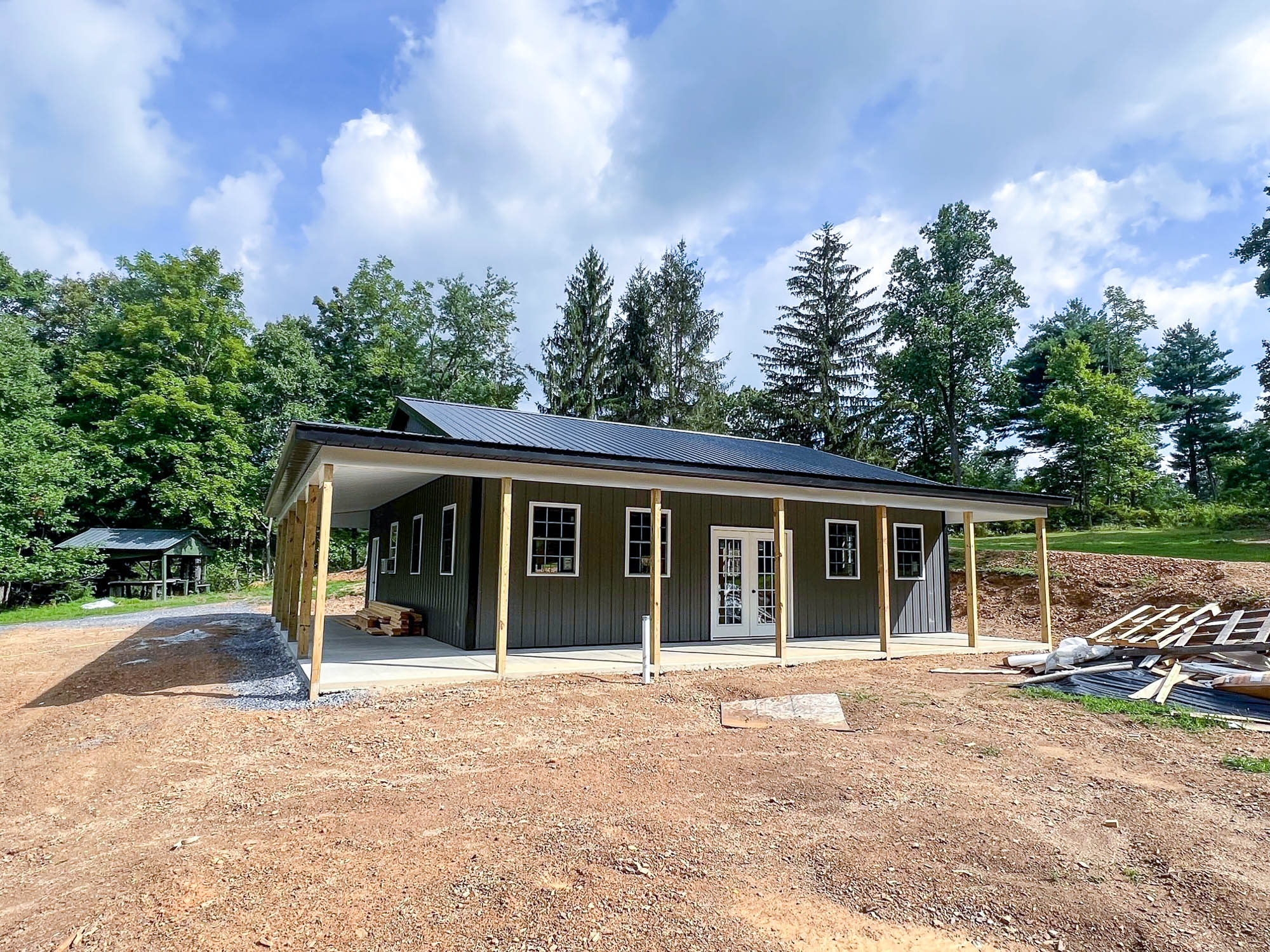 A gray metal building with a gable roof and a covered porch supported by wooden posts. The building is situated on a dirt lot with a wooded area in the background.