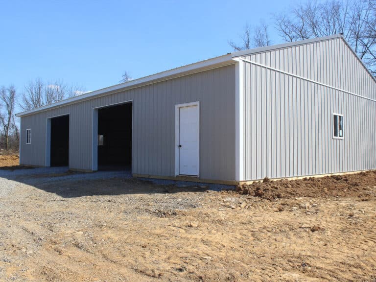 a large gray metal building with dark gray trim, featuring a gable roof and multiple entry points, including a large sliding door and a smaller side door. the structure sits on a dirt lot with construction materials nearby under a clear blue sky.