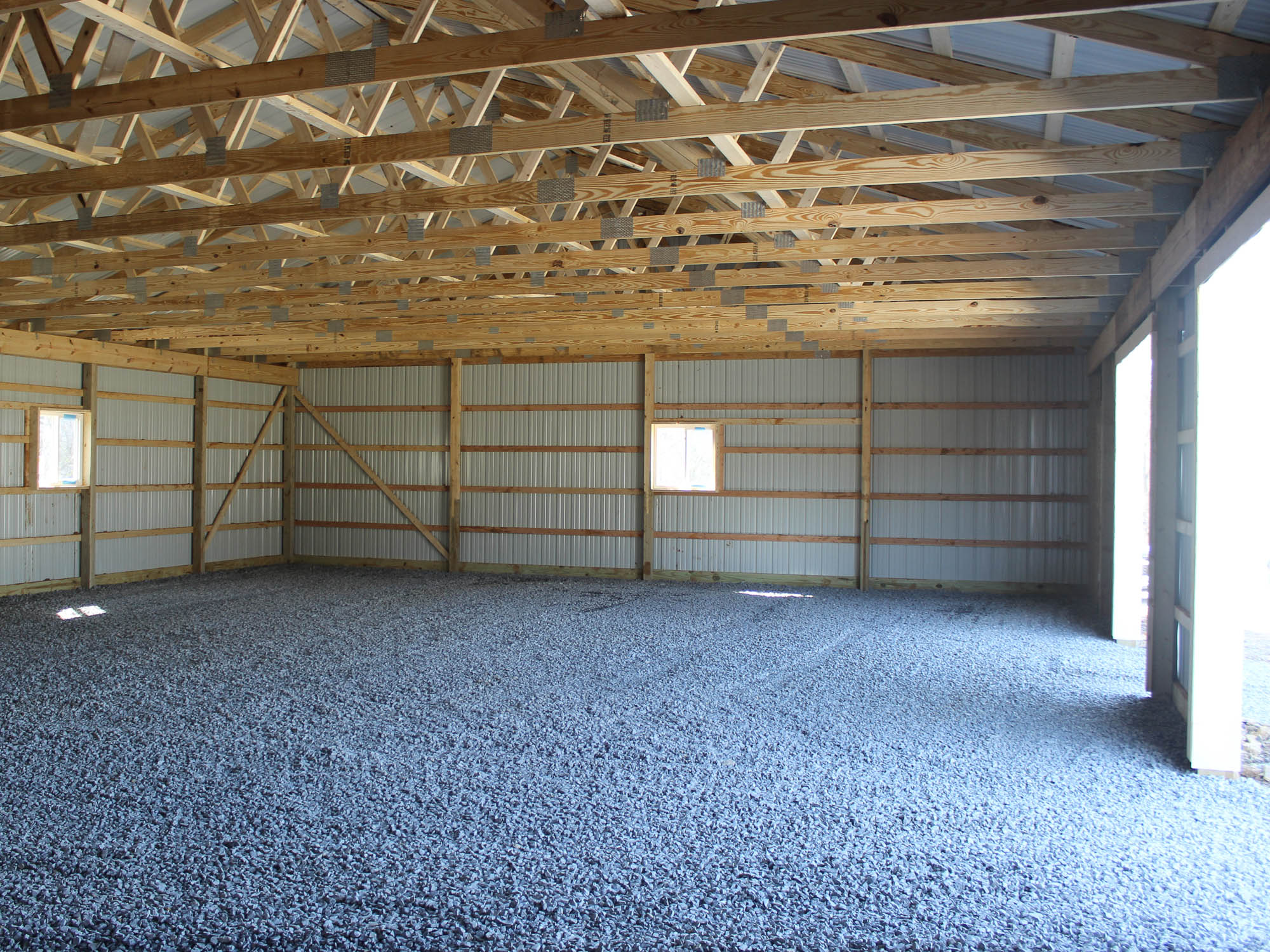 The interior of a metal building with exposed wooden framing, metal siding, and a gravel floor. The ceiling is supported by wooden trusses, and natural light streams in through small windows.