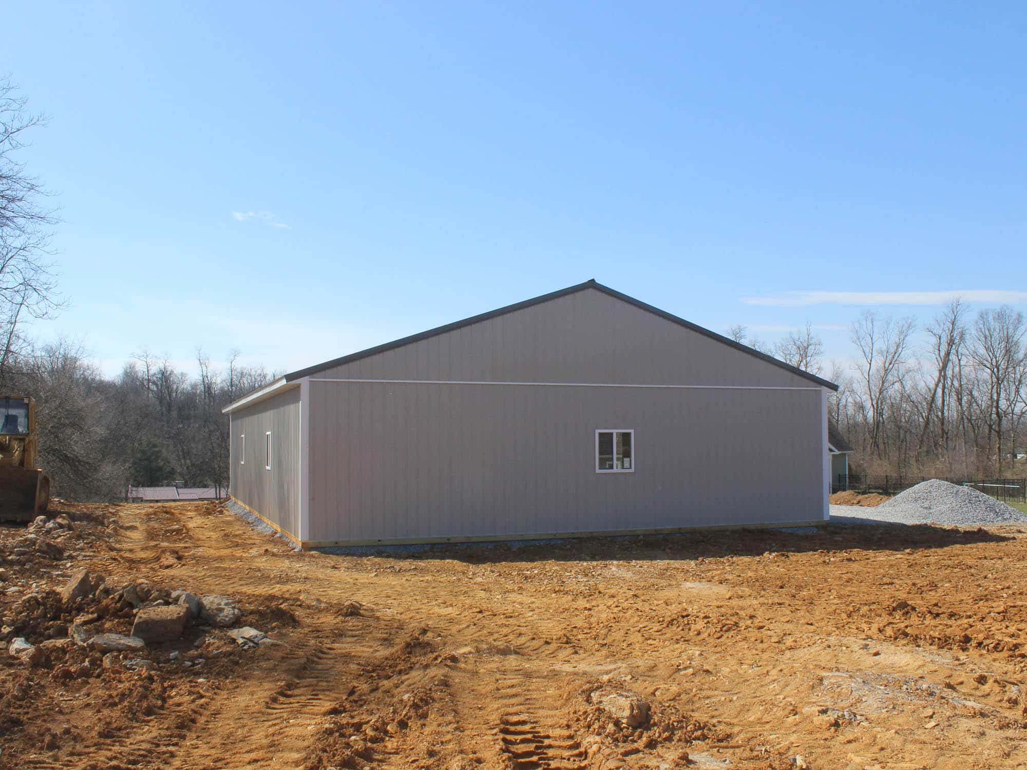 A rear view of a gray metal building with a gable roof and a single small window. The building is surrounded by a dirt lot, with an open sky and trees in the distance.