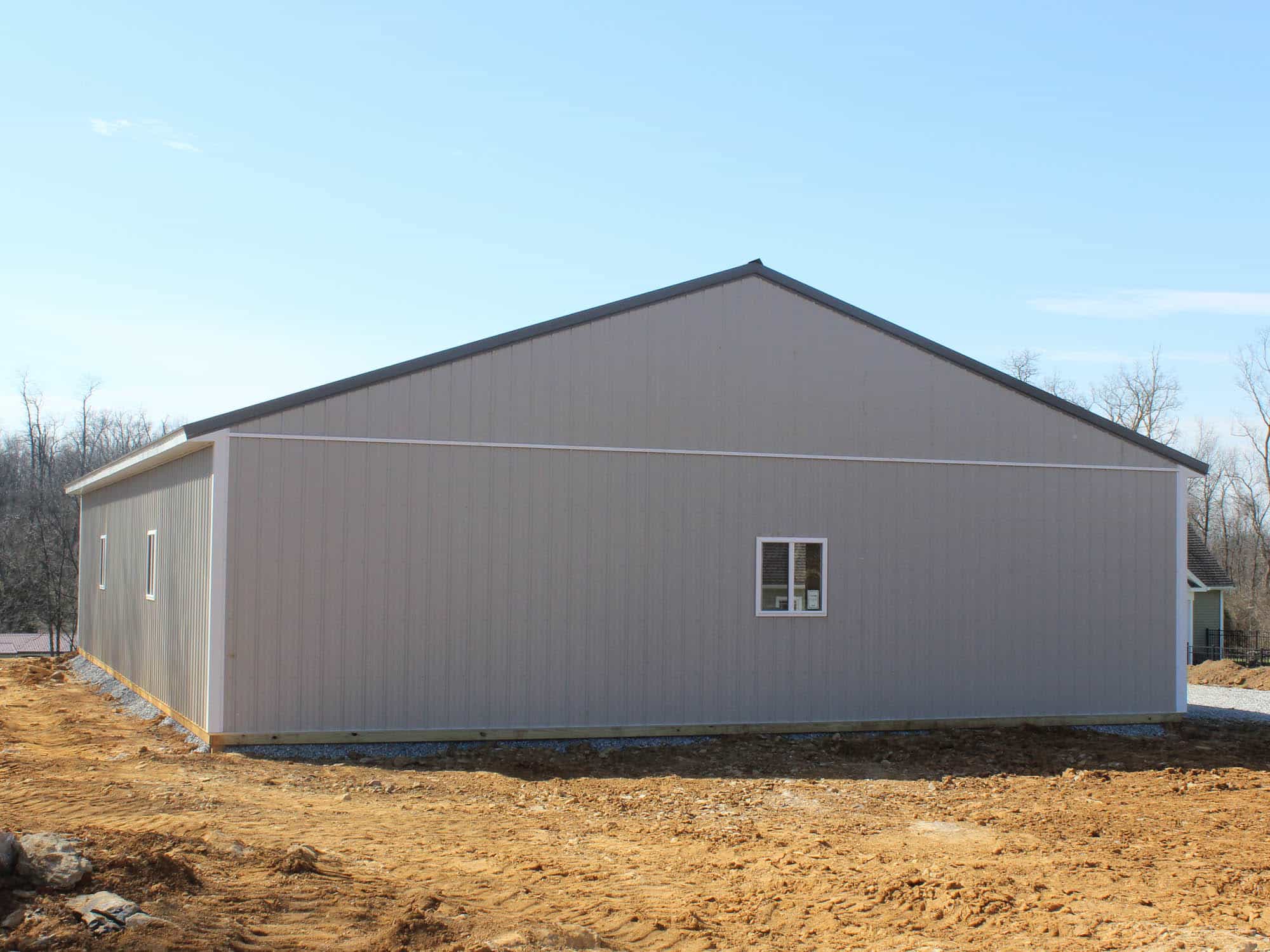 A side view of a gray metal building with a gable roof and a small window centered on the wall. The building is situated on a dirt lot with a clear blue sky and an open landscape in the background.