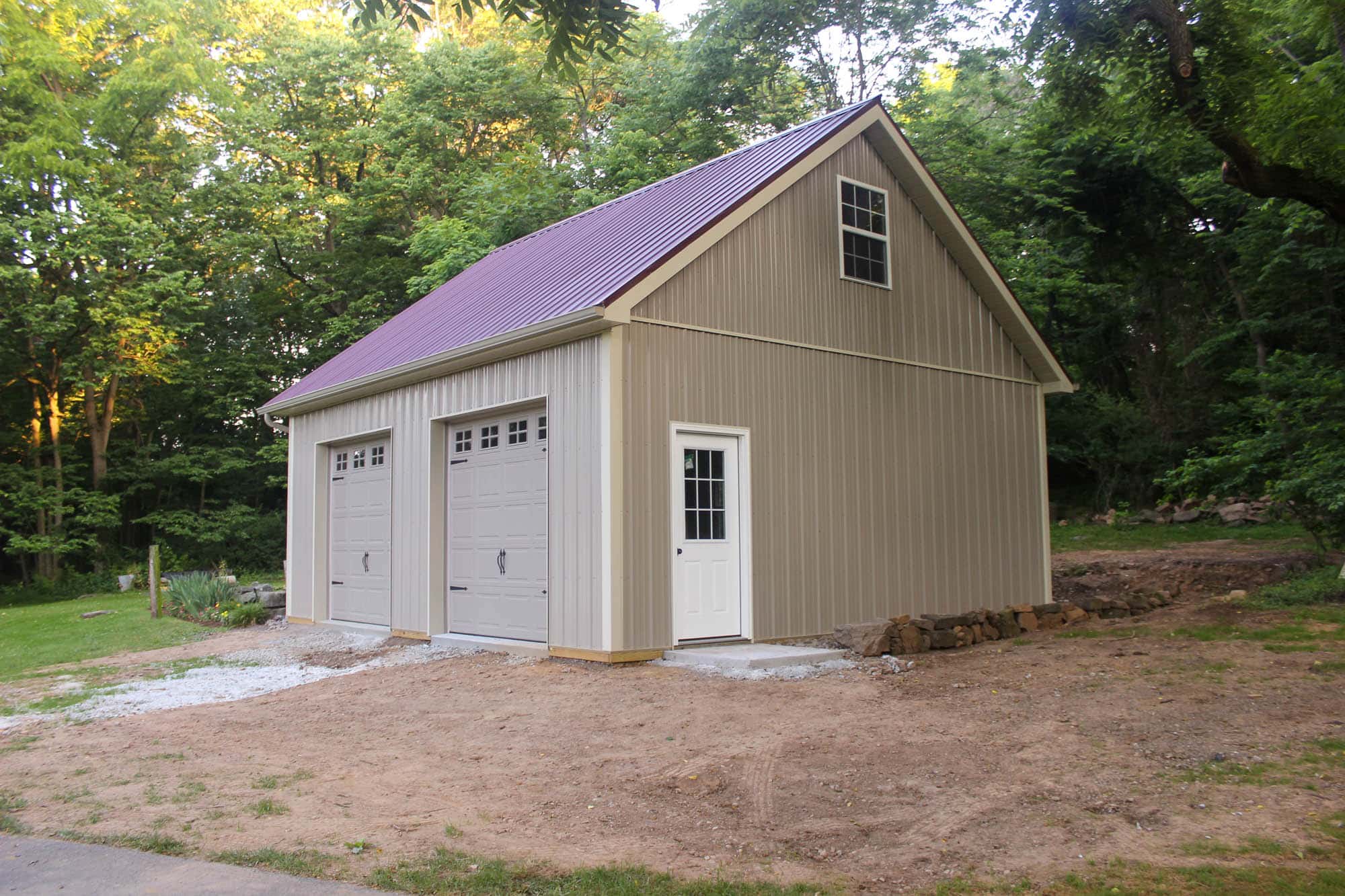 Side-angle view of a beige two-story garage with maroon roof, white trim, and a loft, set among trees with cleared land in front.