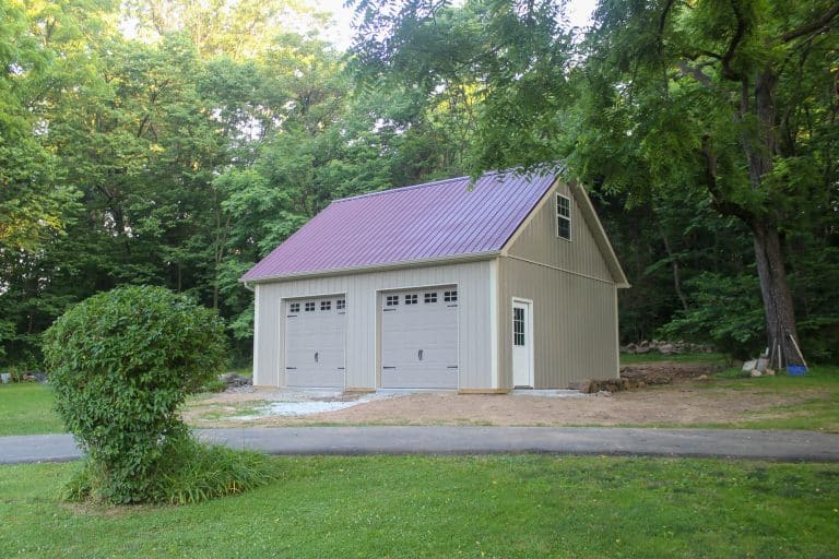 two-story beige garage with maroon roof and white trim, placed on a grassy lot with surrounding trees.