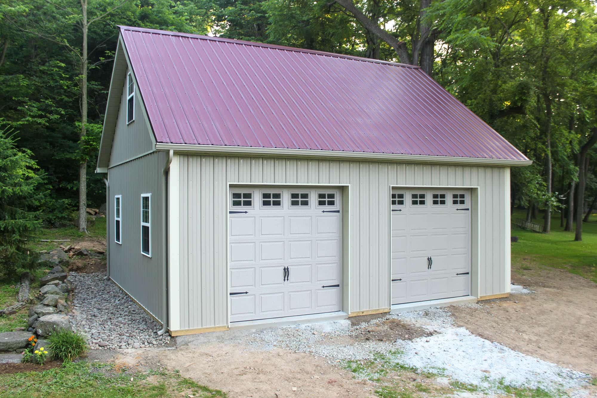 Exterior of a tall two-story garage with dual white doors, beige siding, and a red metal roof, set on a gravel pad.