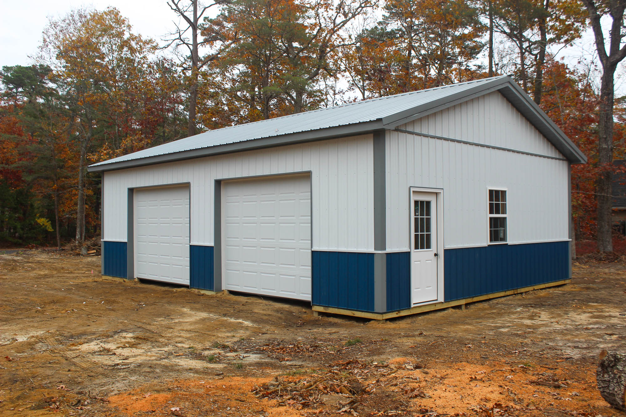 Angled corner view of a blue and white garage building with side windows and overhead doors, situated in a rural area with trees in the background.