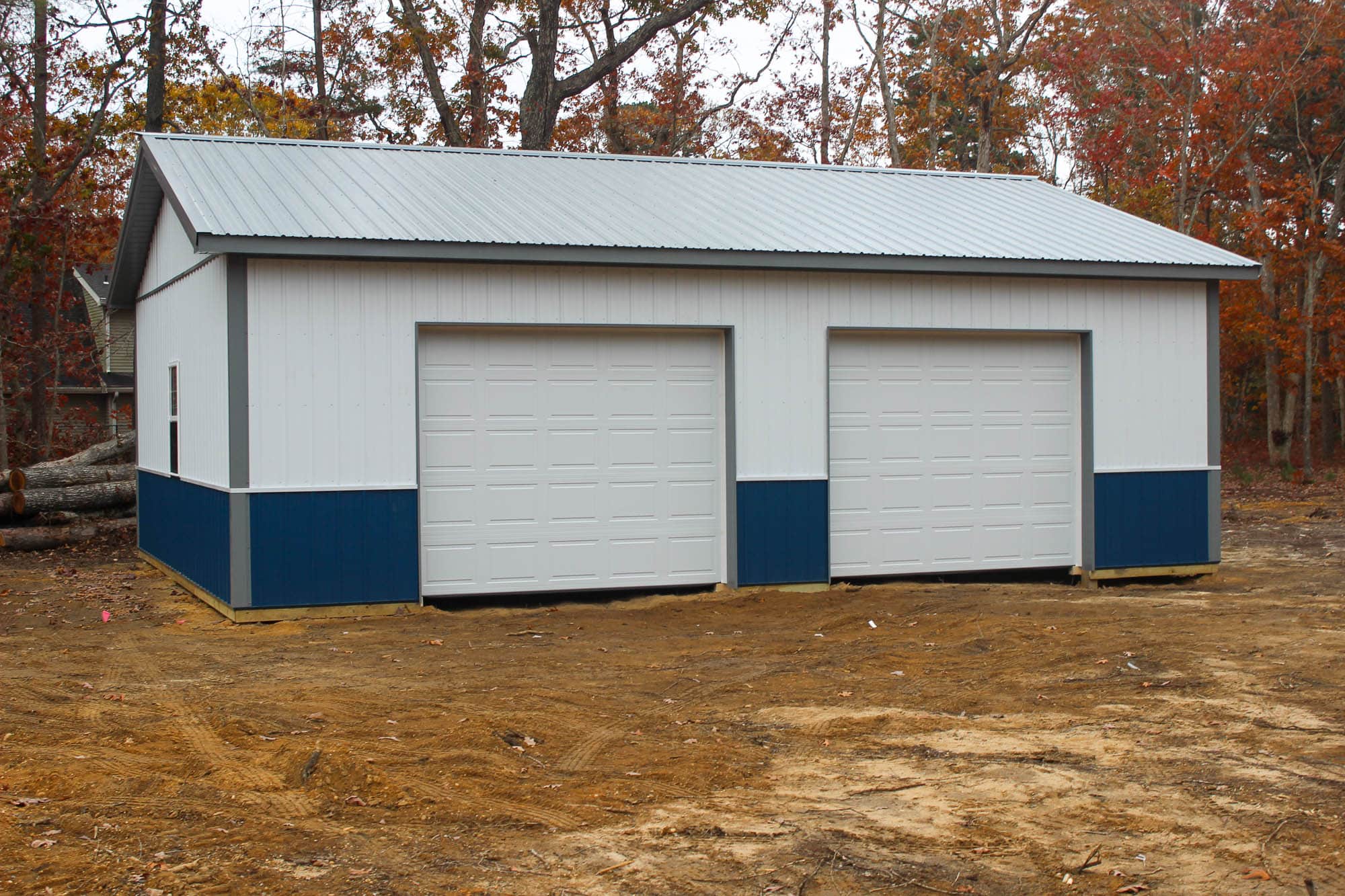 Front view of a metal garage with two large roll-up doors and a central walk-in door, all framed with blue trim, on a cleared dirt lot.