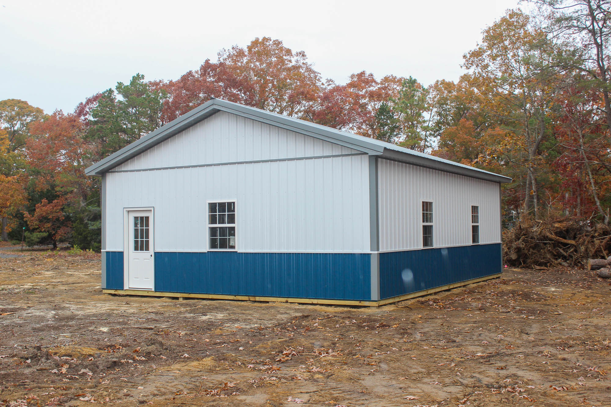 Angled view of a metal garage building with a white exterior, blue base, and walk-in side door, surrounded by autumn trees.