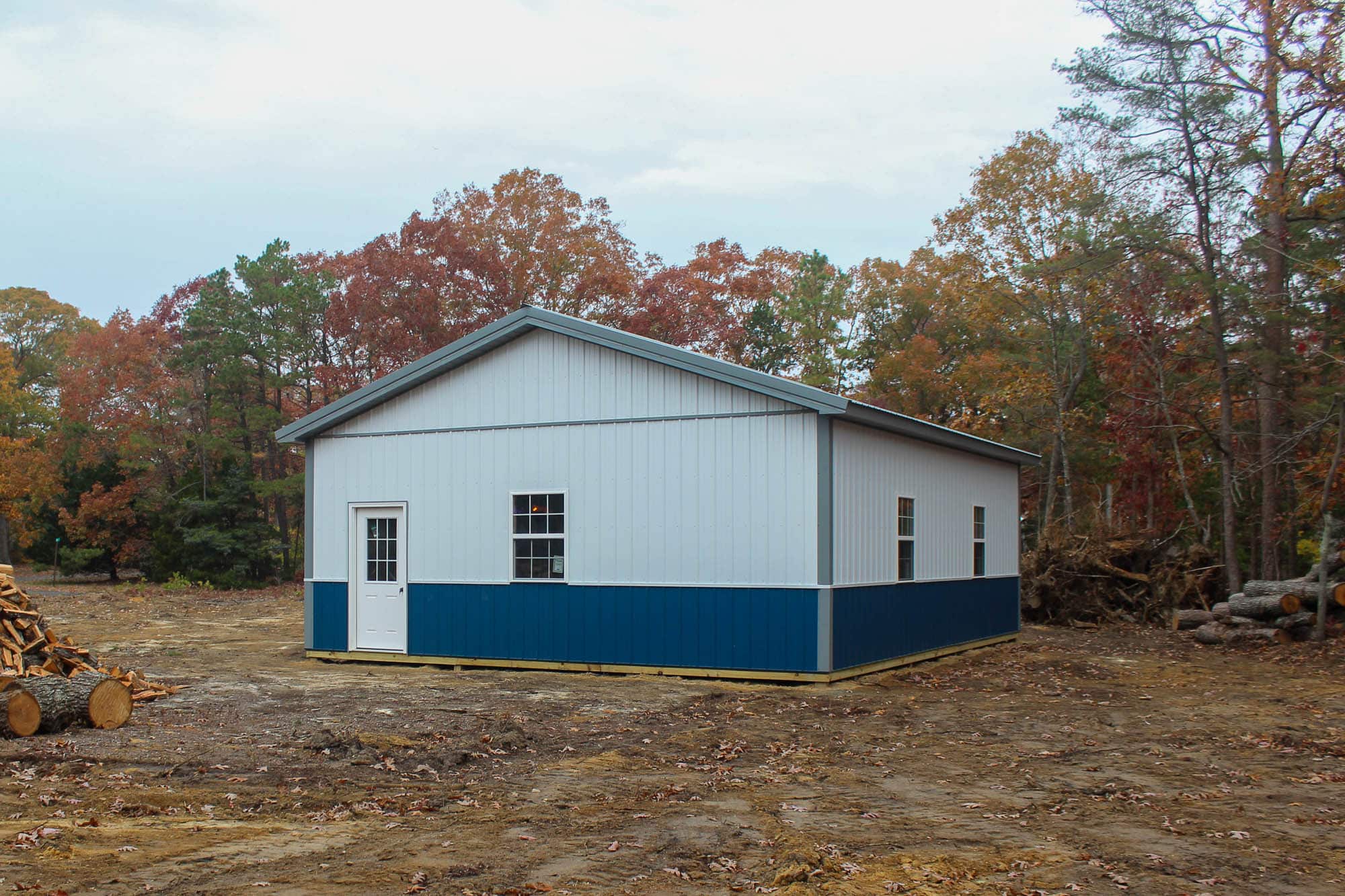 Side view of a blue and white prefab garage with a gable roof and forest backdrop, showing the structure’s clean design and contrasting trim.