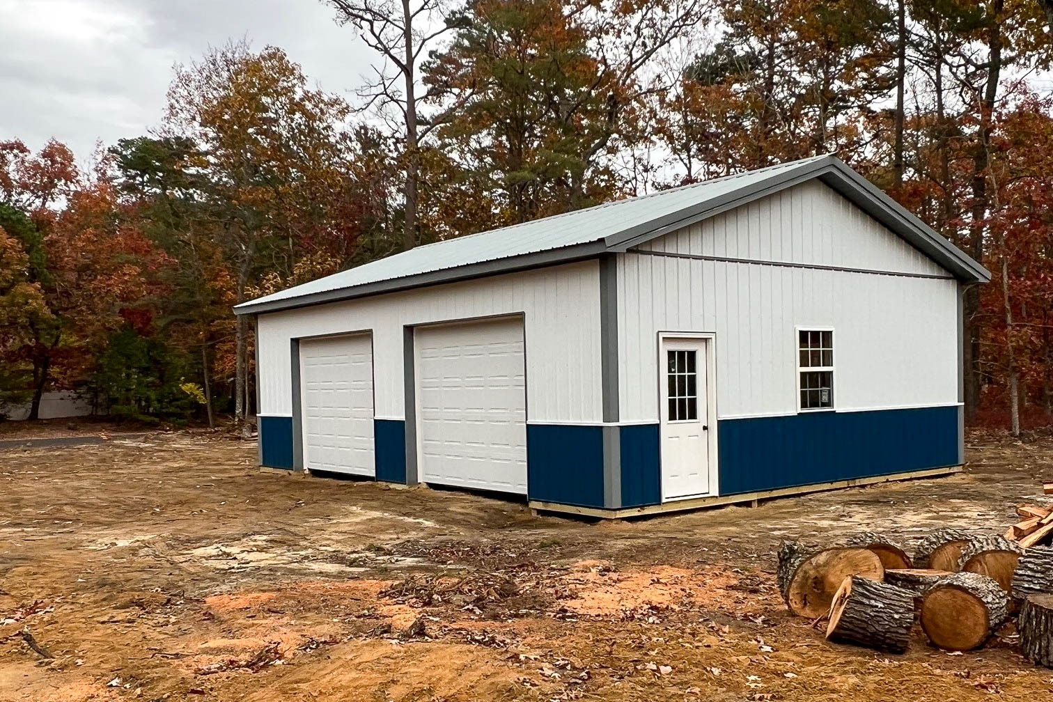 White and blue metal garage with three doors, including two large roll-up garage doors and one walk-in door, set in a wooded area with autumn foliage.