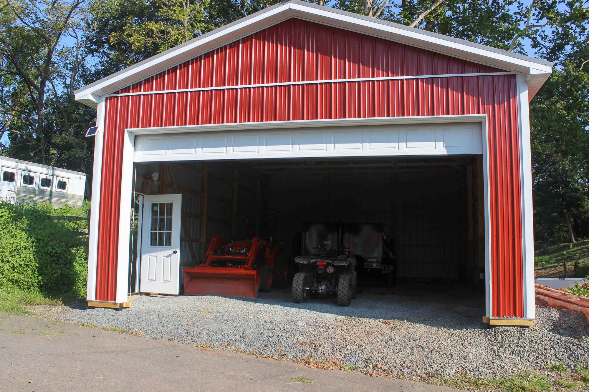 Garage door open to reveal a red metal building with gravel floor interior and lawn equipment inside.