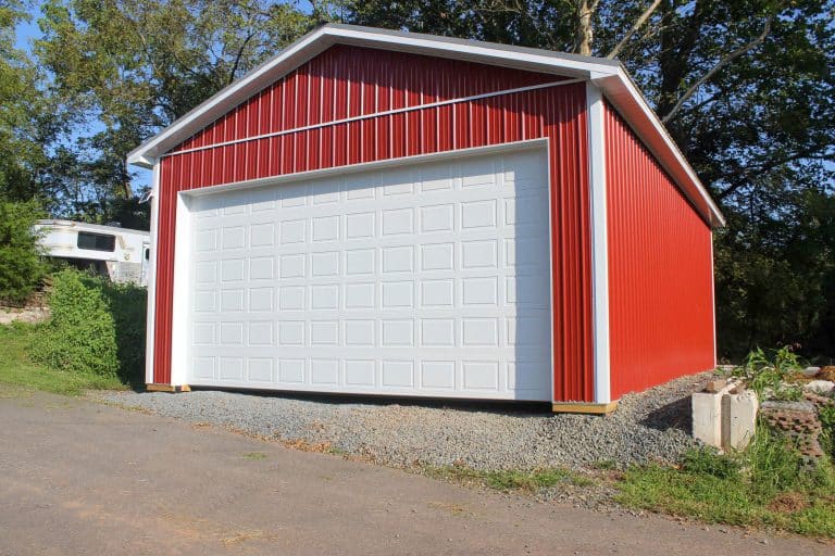 front view of a red garage with a large white roll-up door, situated on a gravel driveway with grass and trees in the background.