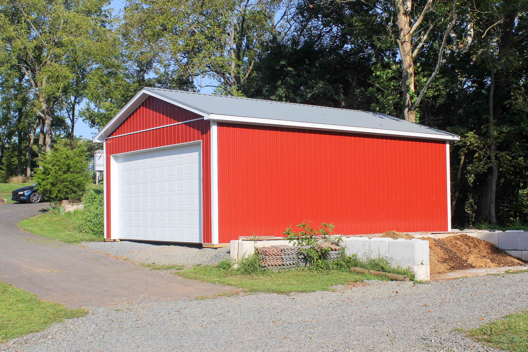 Side view of a red metal garage with a gray roof and white trim, set on a gravel pad beside a wooden pallet.