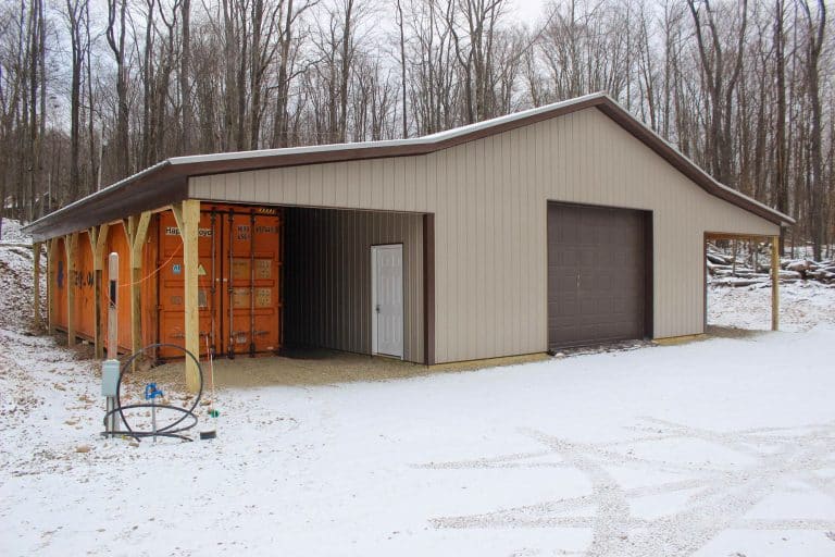 angled view of a two-door garage with a brown overhead door and a covered entry area, surrounded by light snow and dense woods.
