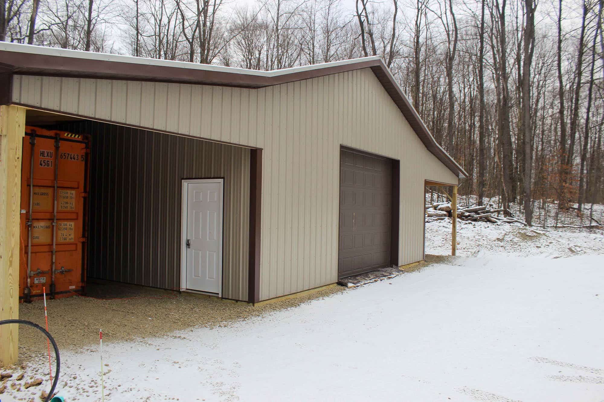 Close side view of a metal garage with a single large roll-up door and walk-in entry, showing brown accent trim and light snow on the ground.