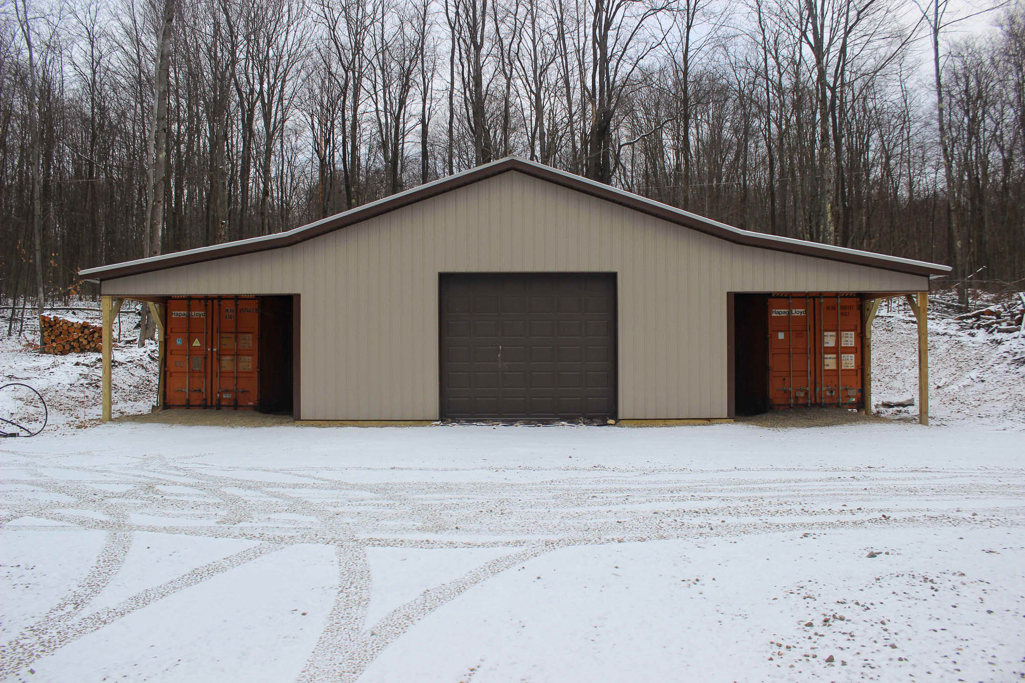 Wide metal garage with tan siding, dark brown trim, and two roll-up doors, surrounded by snow-covered ground and bare trees in the background.