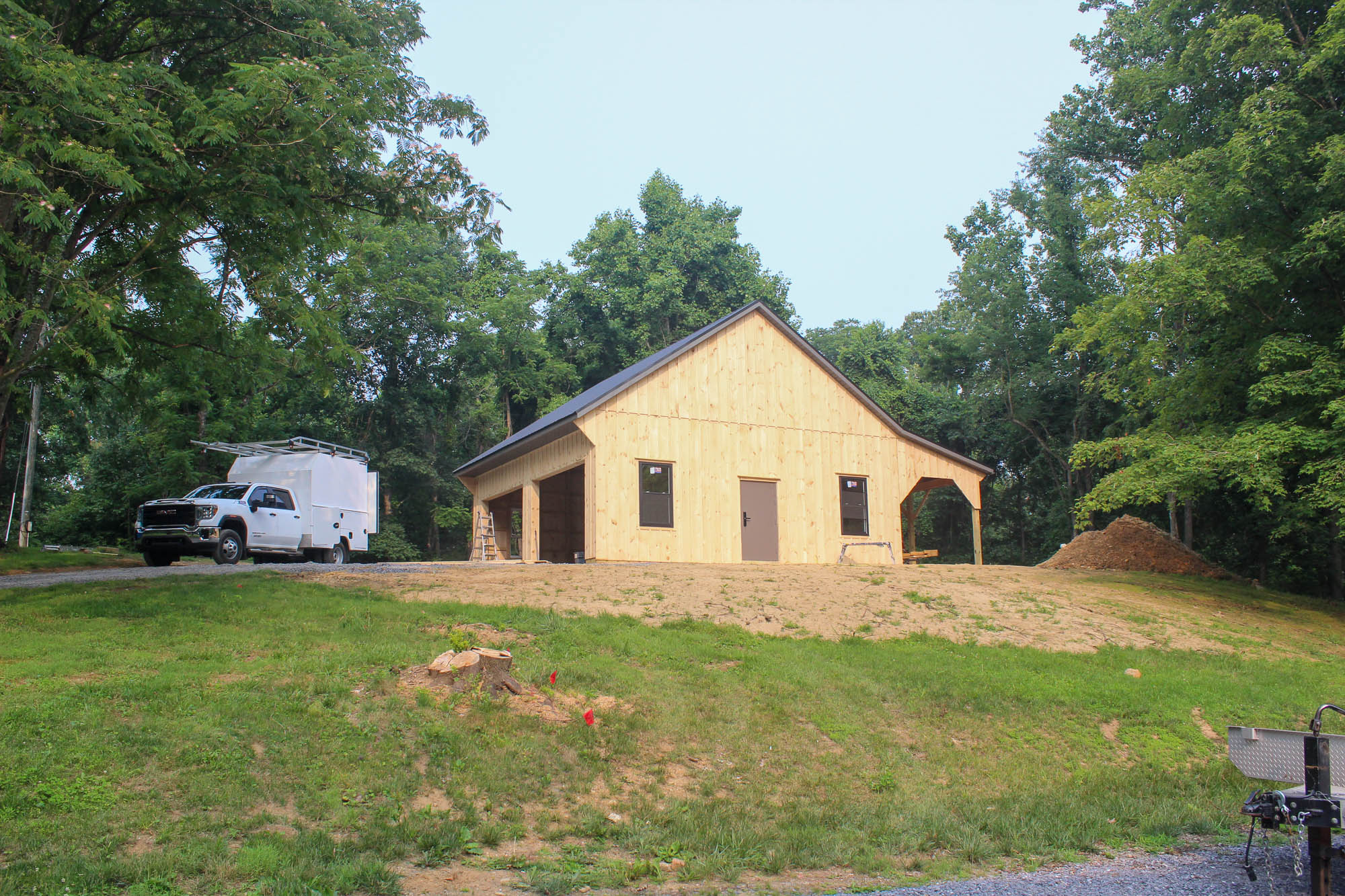 Newly constructed wooden garage or shop with light vertical board siding, dark trim, and a black metal roof, located at a wooded property with a white utility truck parked nearby.
