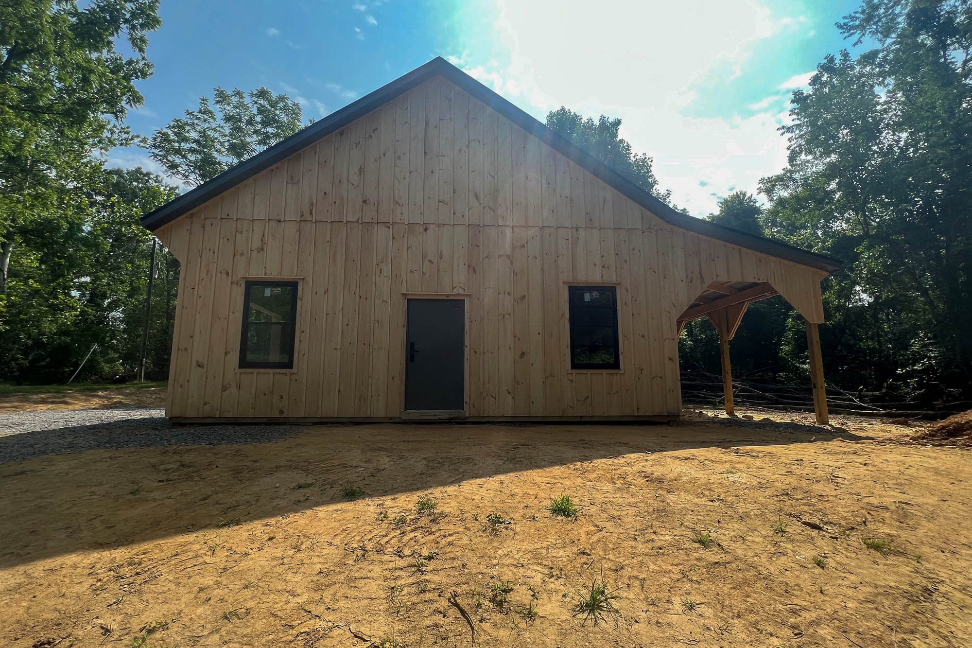 Front view of a custom wooden garage with twin black garage doors and a gabled roof, surrounded by trees and set on a dirt lot.