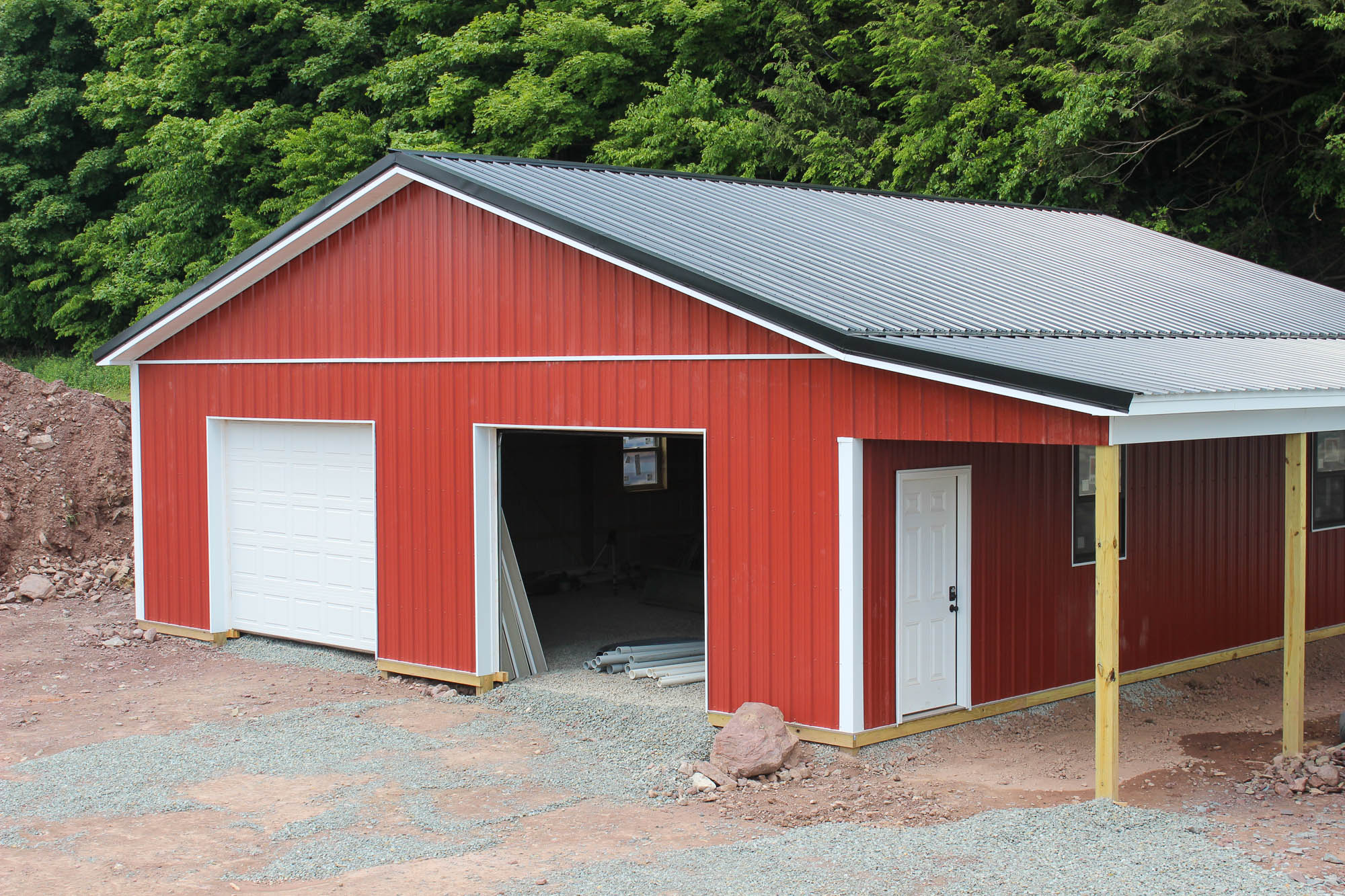 Front and side view of a red double-door garage with open bays and lean-to roofing, partially completed with gravel in front.