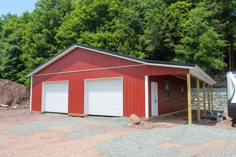 angled view of a red garage with dual white garage doors and a side lean-to under construction, surrounded by forest.