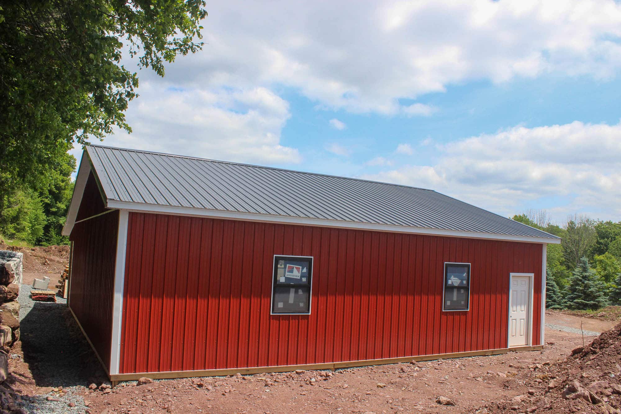 Side view of a red metal garage with dark roof and single window, resting on a gravel lot beneath a partly cloudy sky.