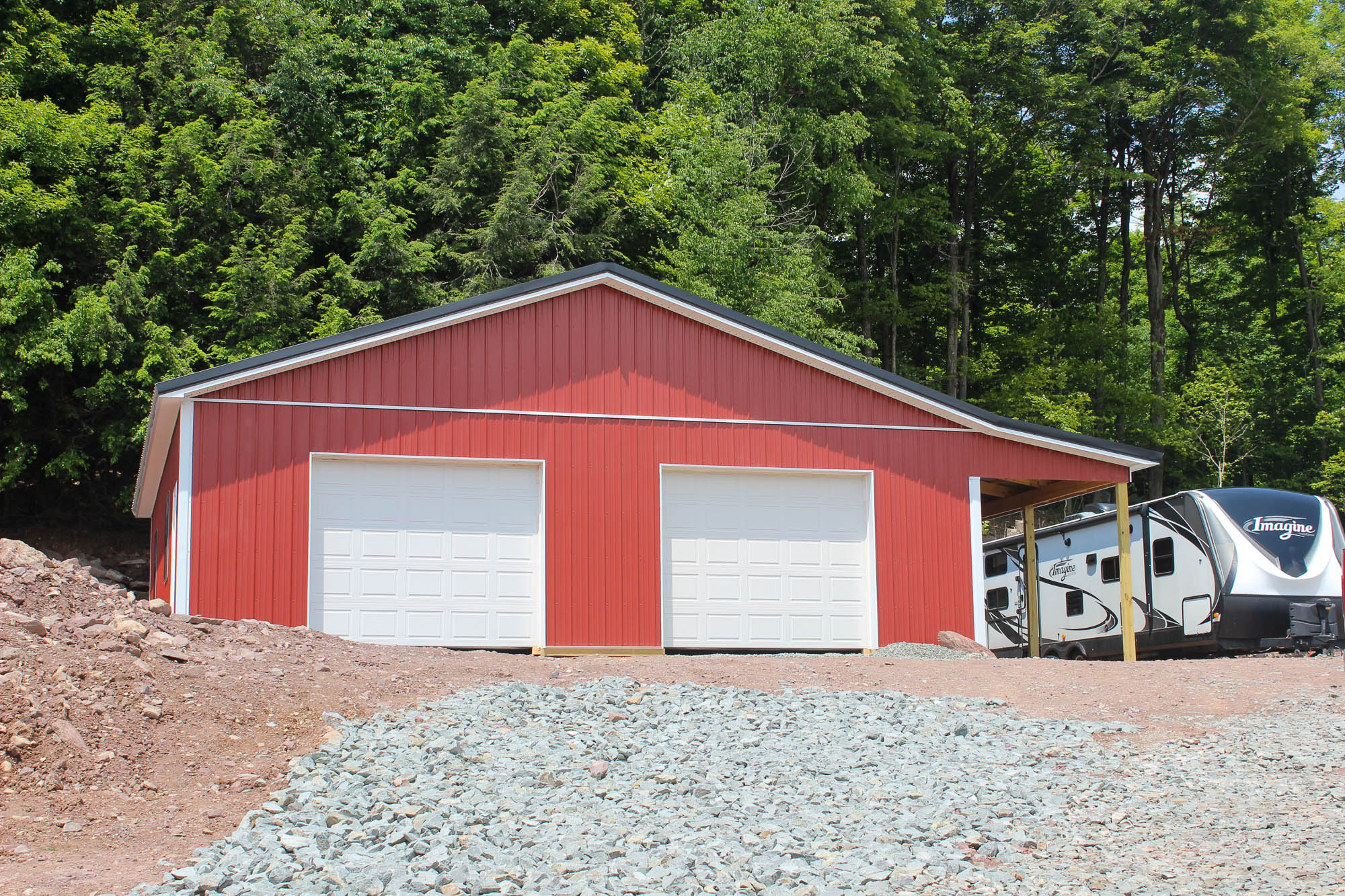 Front view of a finished red garage with dual white overhead doors, gravel driveway, and surrounded by trees and vehicles.