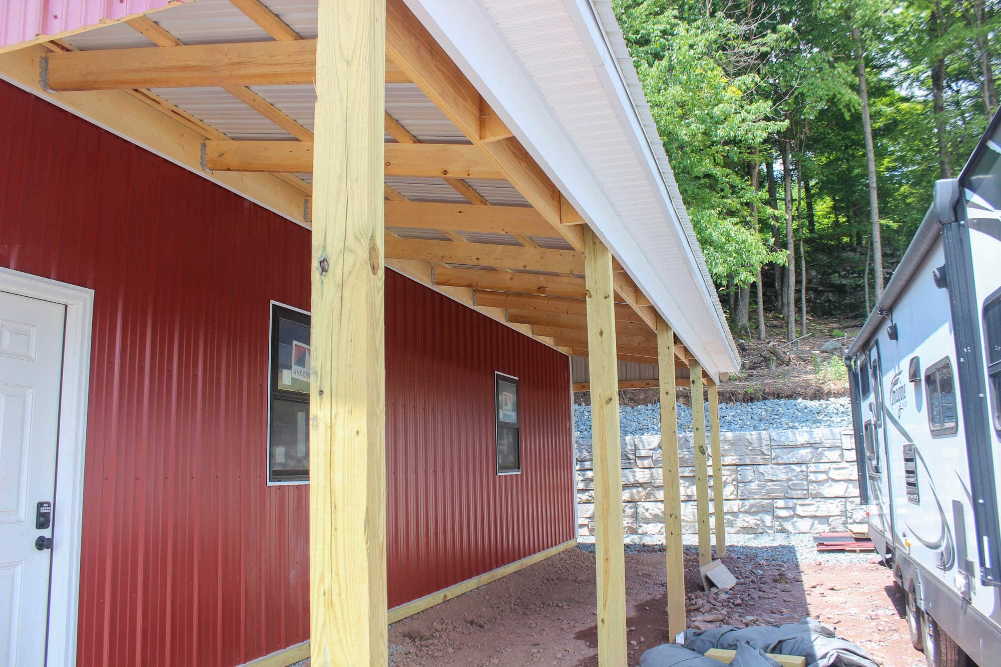 Close-up view of a red garage's side porch, featuring vertical metal siding, wood posts, and gravel foundation with construction equipment nearby.