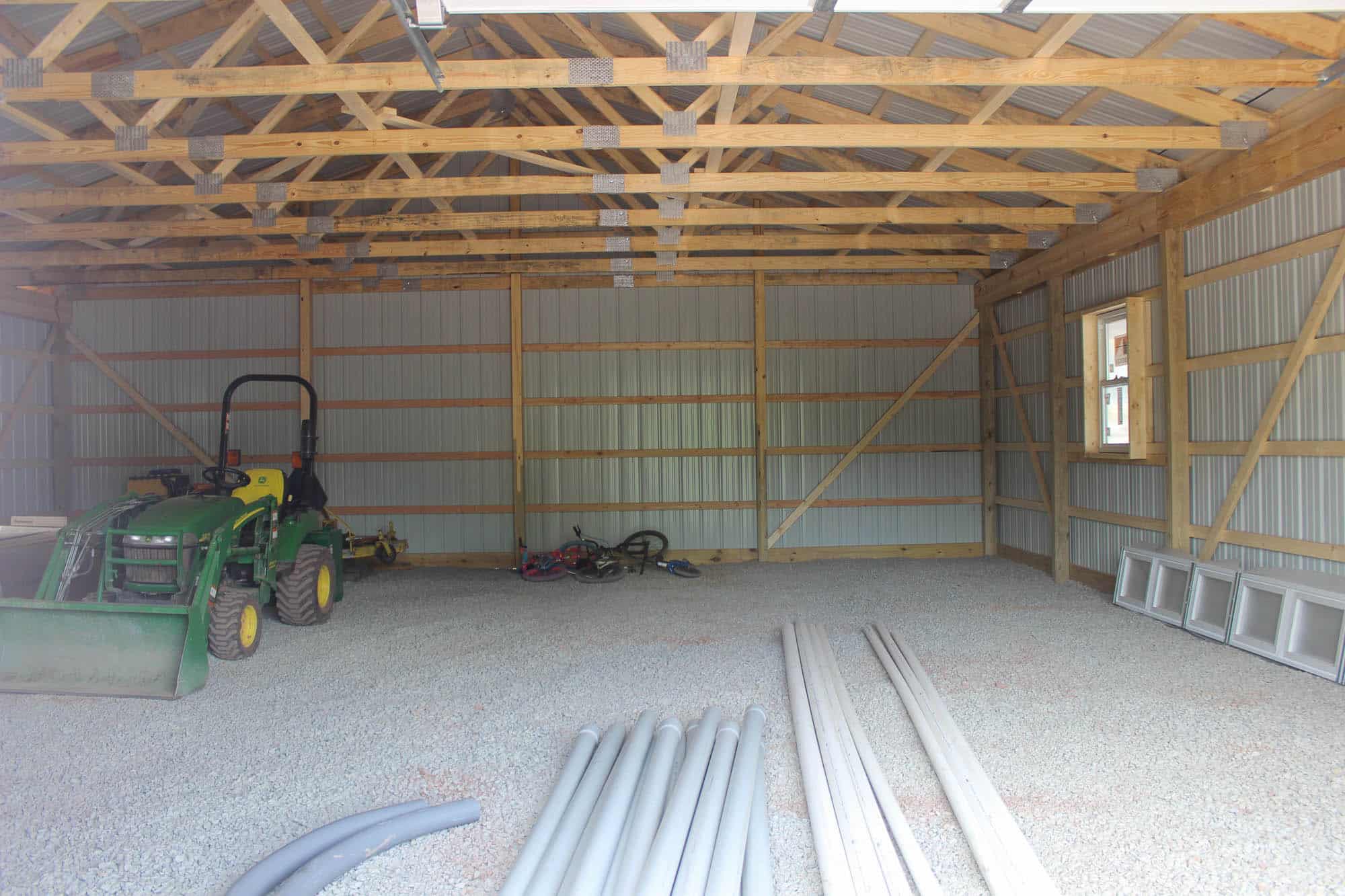 Interior view of a red prefab garage under construction, showing open framing, wooden trusses, and construction materials laid out on the concrete floor.