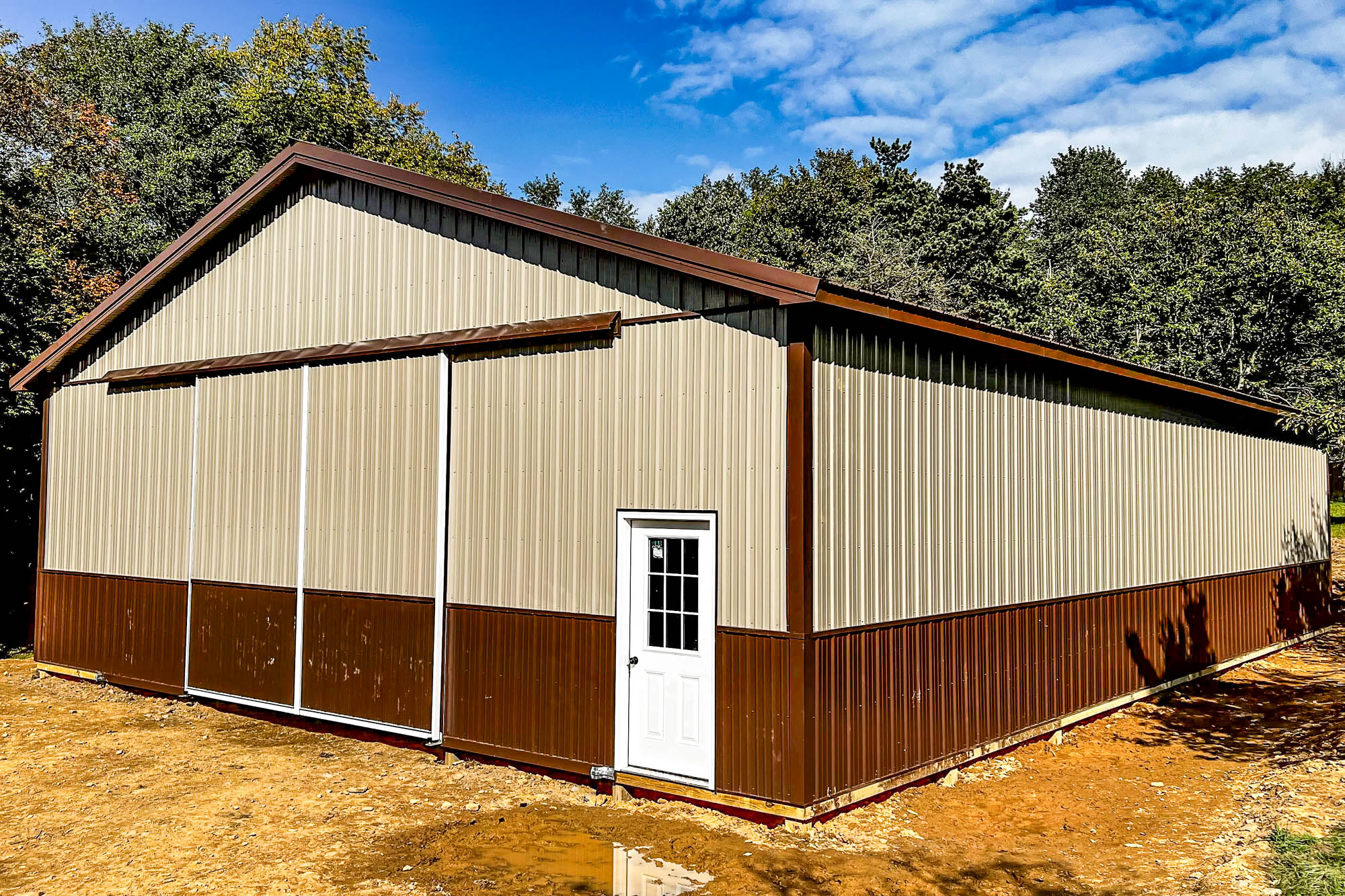 Side view of a tan-and-brown steel warehouse with a white entry door and large sliding doors, placed on a cleared construction site surrounded by forest.