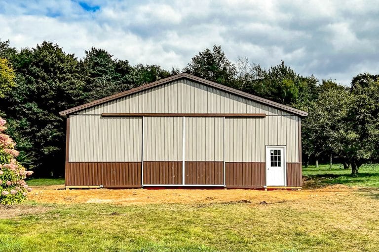 a large tan-and-brown metal warehouse building with a wide overhead door and a single side entry door, situated on a dirt lot with trees in the background.