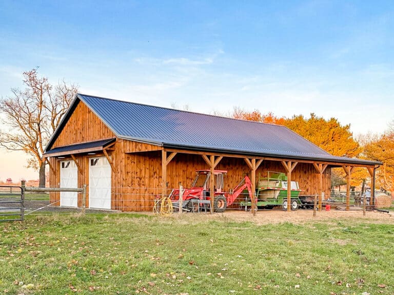 a wooden pole barn with an open side, featuring a dark metal gable roof and natural wood siding. the open section provides easy access to farm equipment, hay storage, or livestock shelter. the barn is surrounded by a grassy field with a clear sky at sunset. learn more about new holland supply's pole barn kits.
