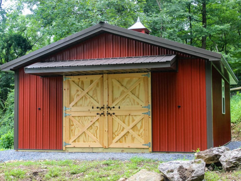 a traditional red pole barn with a dark metal roof, featuring large wooden double doors with an x-brace design. a cupola sits atop the roof, and the barn is surrounded by greenery and a gravel pathway leading to the entrance.