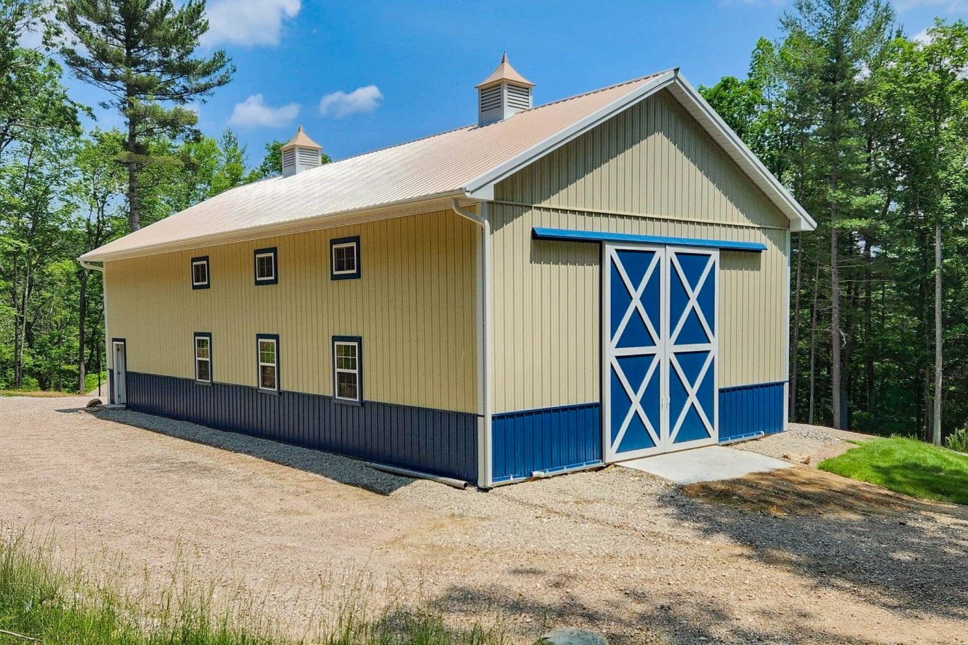 beige and blue pole barn with large sliding barn doors, metal roofing, cupolas, and gravel access area.