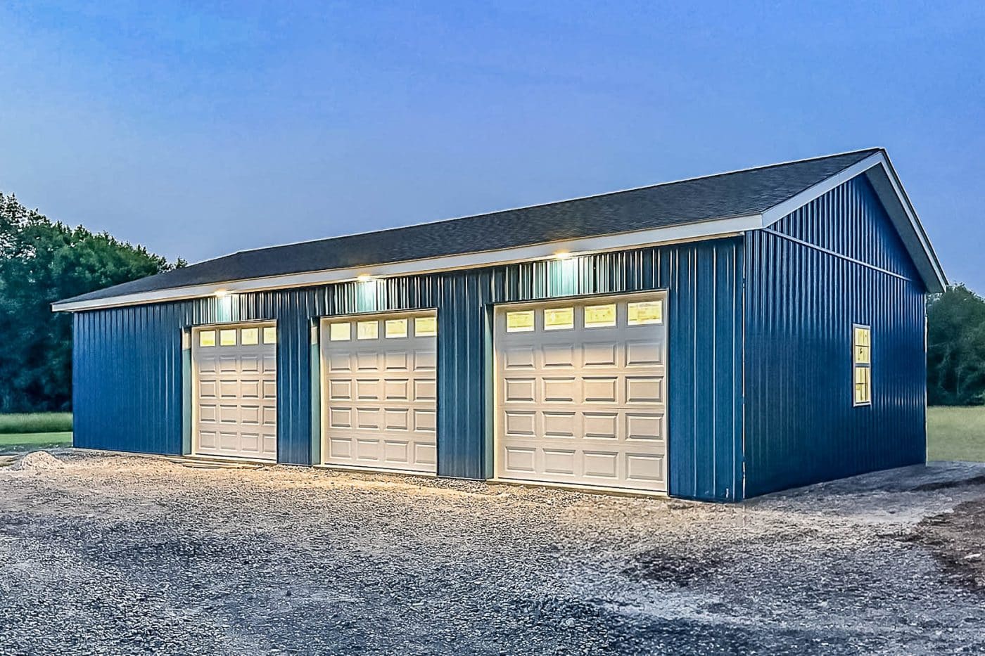blue pole garage with three white overhead doors and exterior lights at dusk.