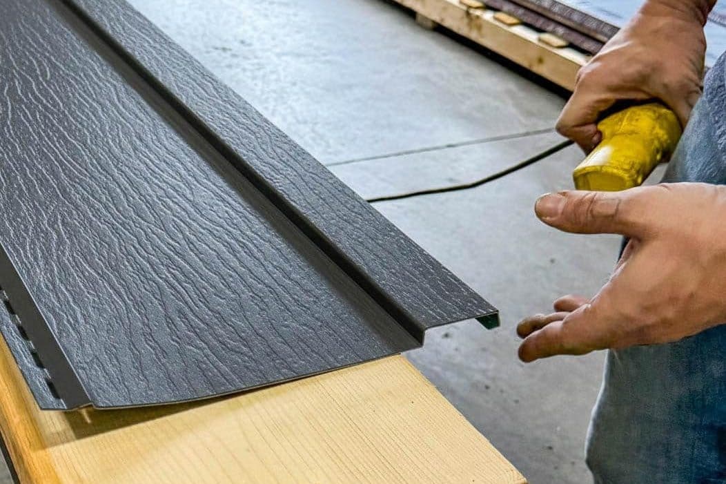 worker handling dark gray board and batten metal siding panel with embossed wood-grain texture on a workbench.