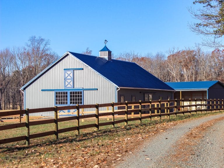 gray pole barn with blue trim and roof, fenced in with wooden rails, on a clear day in new york.