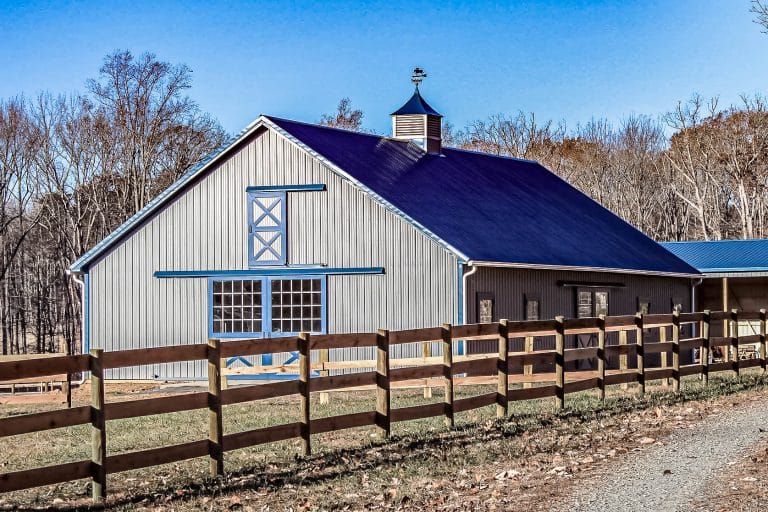 gray pole barn with blue trim, cupola, large doors, and wooden fence.