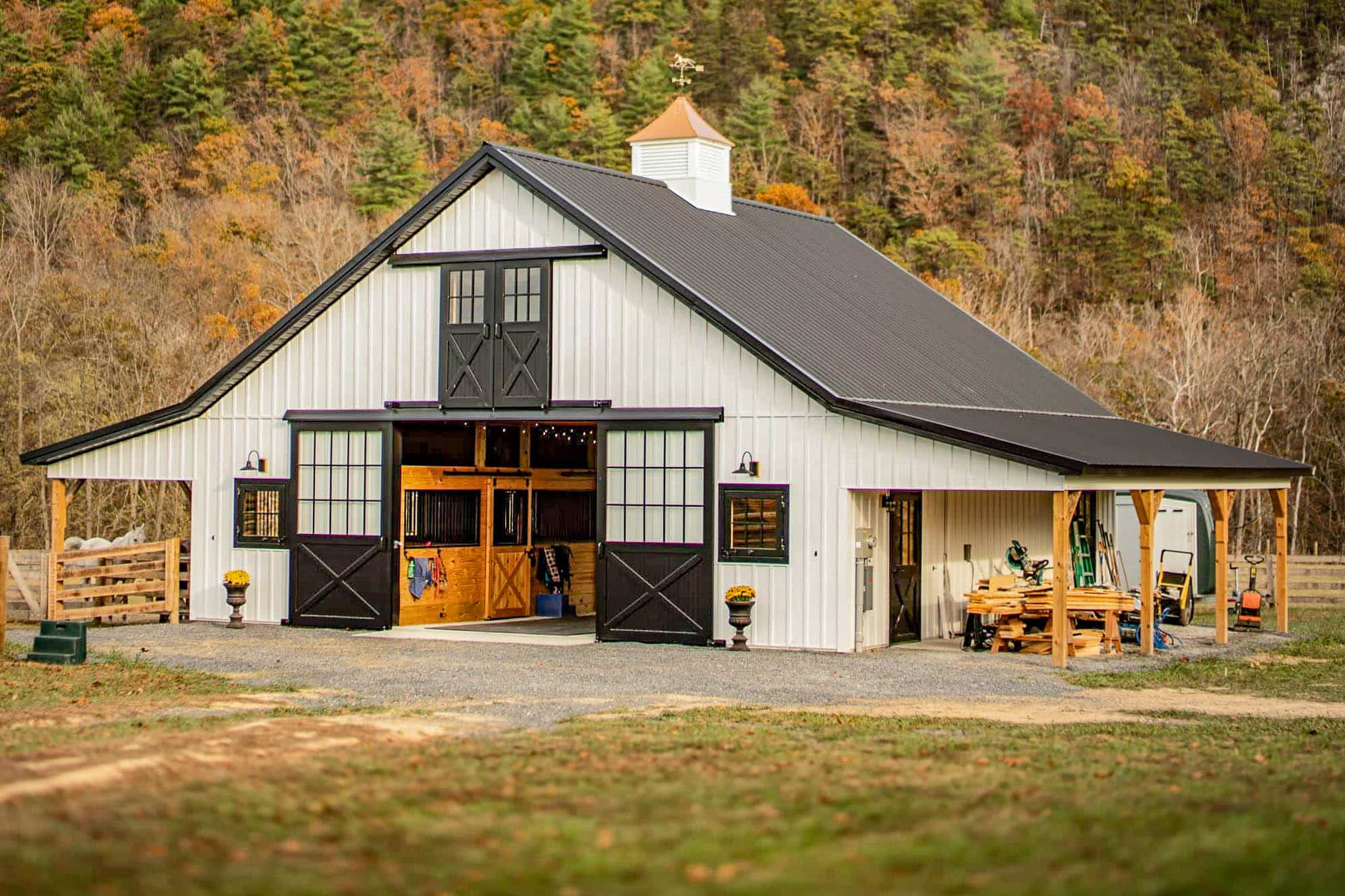 white horse barn with black trim, open center entry, dutch doors, cupola, and attached covered work area.