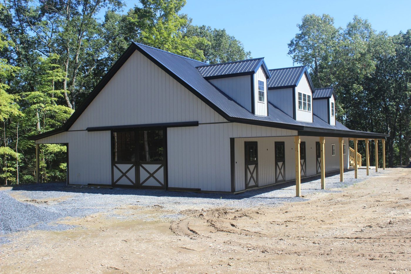 large white post-frame horse barn with black metal roof, dormers, sliding doors, covered porch, and gravel entrance.