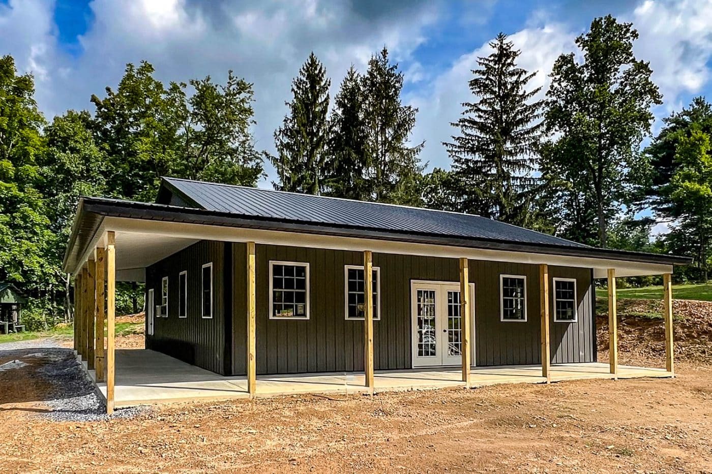 dark post-frame building with wraparound porch, wood posts, metal roof, windows, and double entry doors.