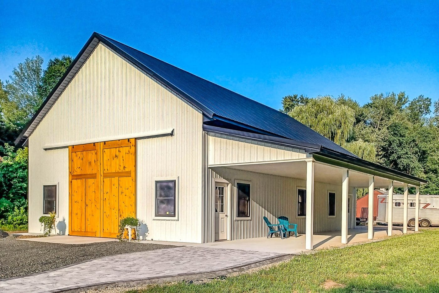 beige pole barn with black metal roof, large wooden sliding doors, covered porch, and concrete entry area.