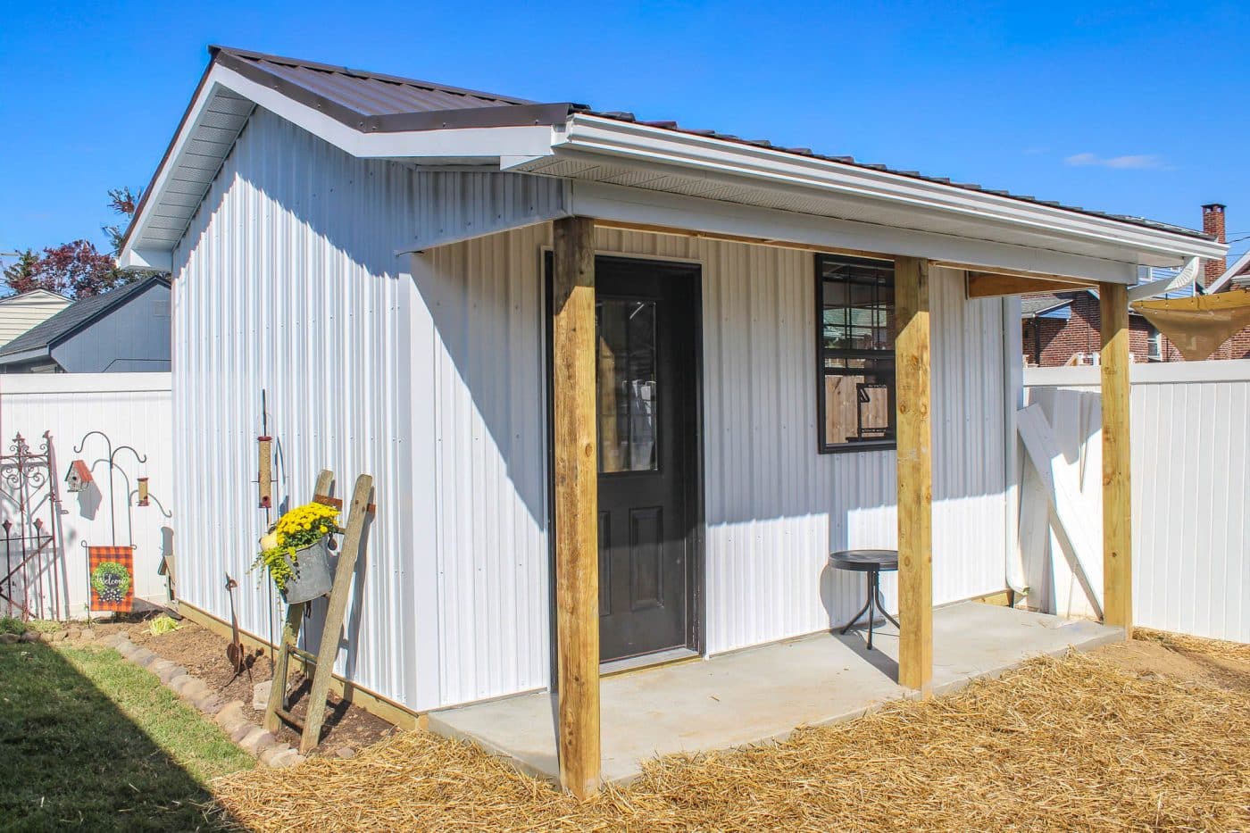 white post-frame shed with metal siding, covered porch, wood posts, black entry door, and concrete walkway.
