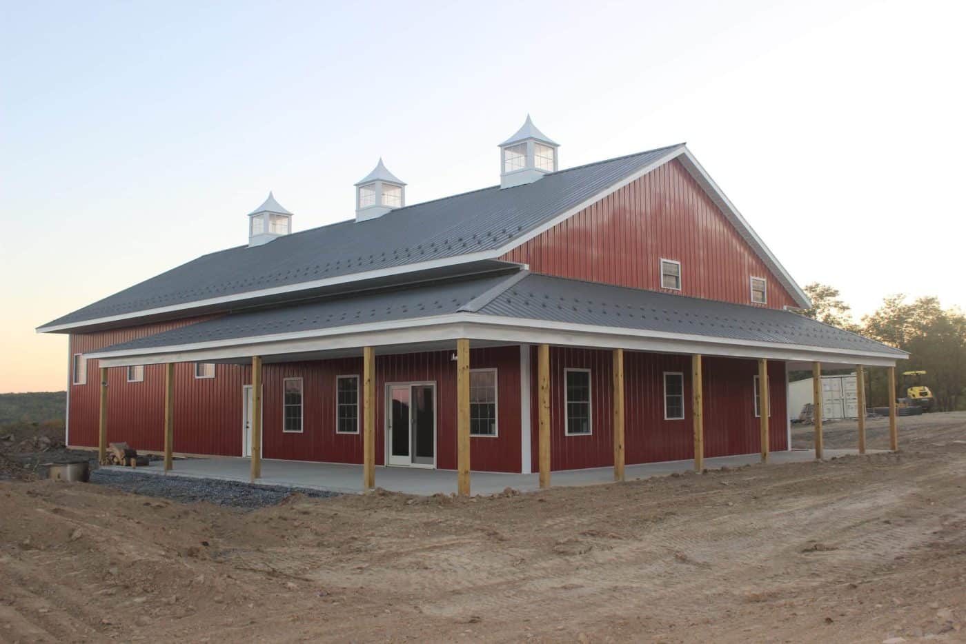 red and white post-frame barn with a wraparound porch and three roof cupolas under a clear sky.
