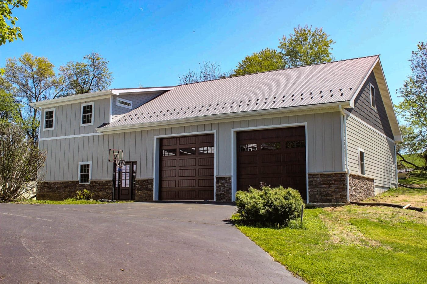 large beige post-frame garage with two brown overhead doors, stone accents, metal roof, and basketball hoop.