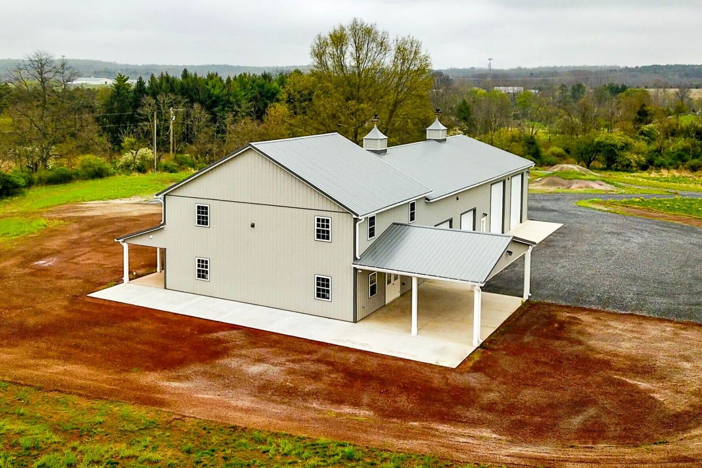 aerial view of a tan post-frame barndominium with metal roof, cupolas, covered porch, gravel driveway, and rural property.