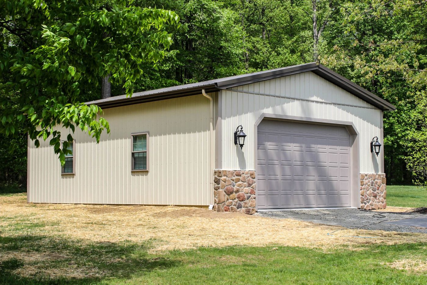 tan post-frame garage with stone accents, overhead door, metal siding, side windows, and wooded background.
